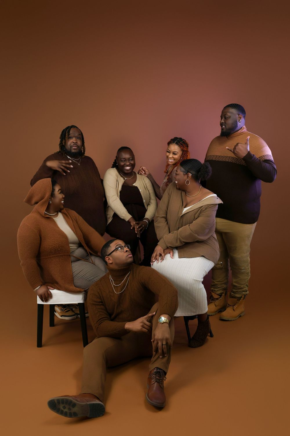 A group of six individuals poses together in a studio setting against a brown backdrop wearing earth-toned clothing.