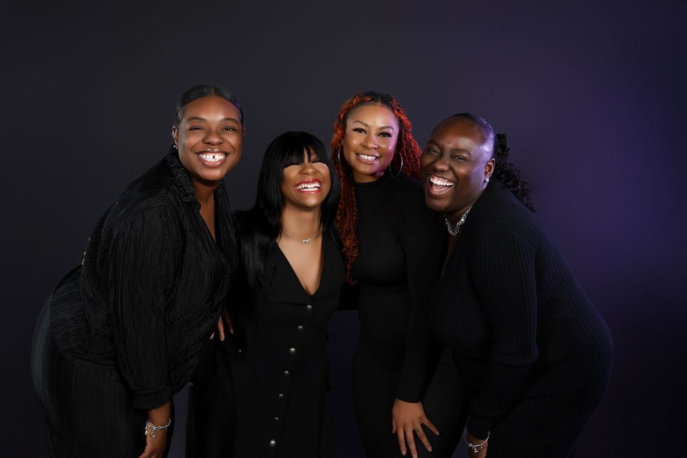 A joyful group of friends wearing black outfits pose together against a dark background with dramatic lighting.