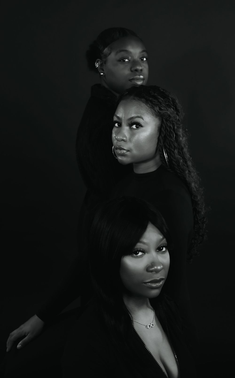Black and white artistic portrait of three women wearing black outfits posed dramatically against dark background.