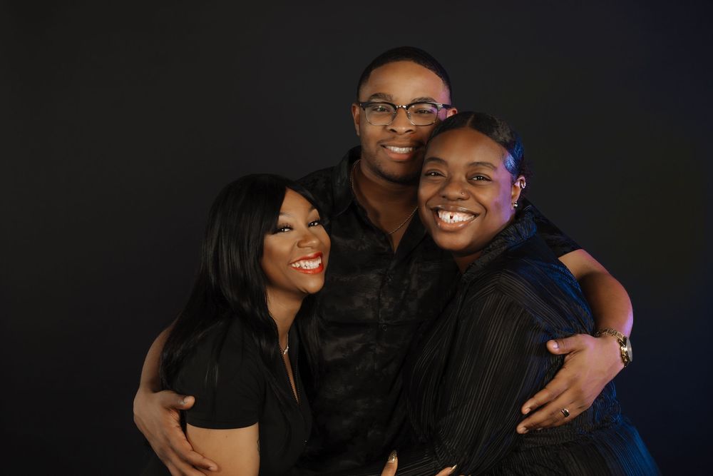 A close-up portrait of three people embracing and smiling warmly against a dark background.