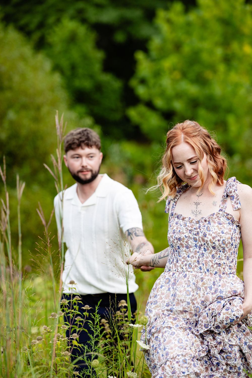 Redheaded woman in floral dress holding hands with tattooed bearded man walking through lush green field.
