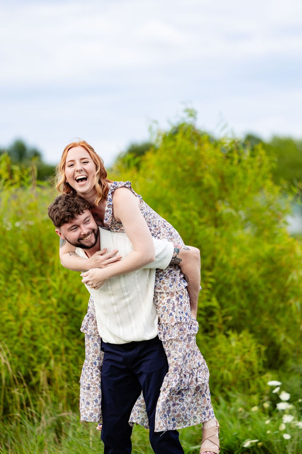 Laughing woman in floral dress getting a piggyback ride from smiling bearded man in green outdoor setting.