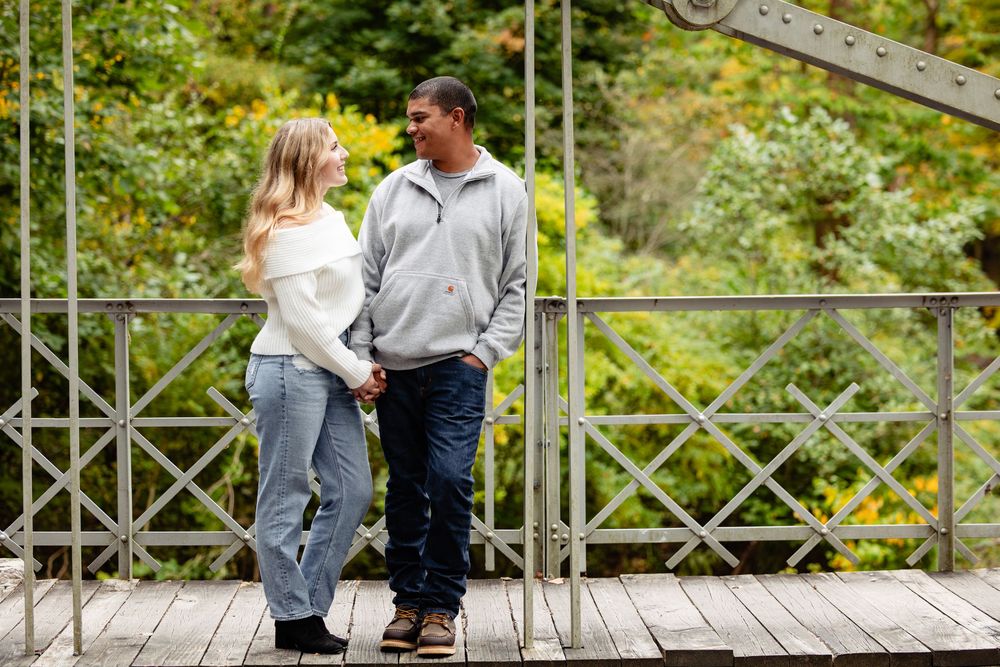 Interracial couple holding hands and gazing at each other on a metal bridge surrounded by lush green foliage.