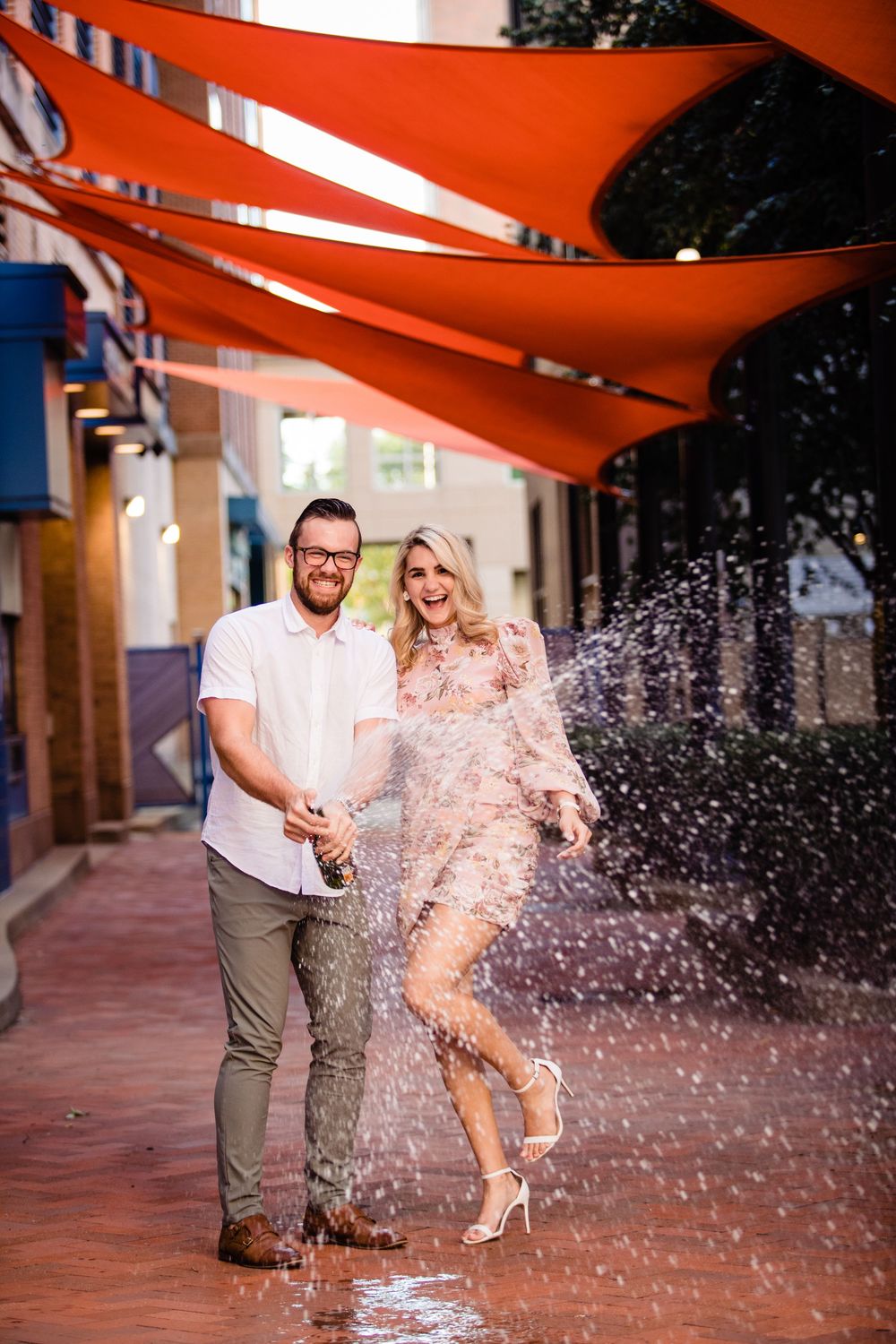 Couple celebrating with champagne spray on a colorful urban street under red canopies.