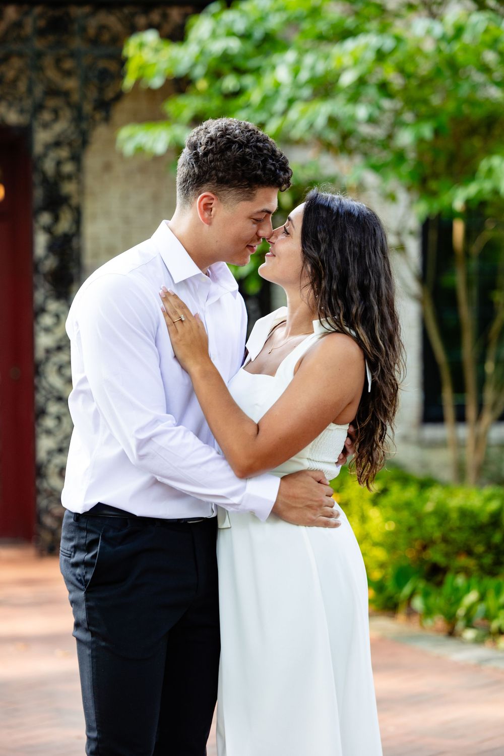 Couple in white outfits sharing a romantic nose-to-nose moment in a tropical garden courtyard setting.