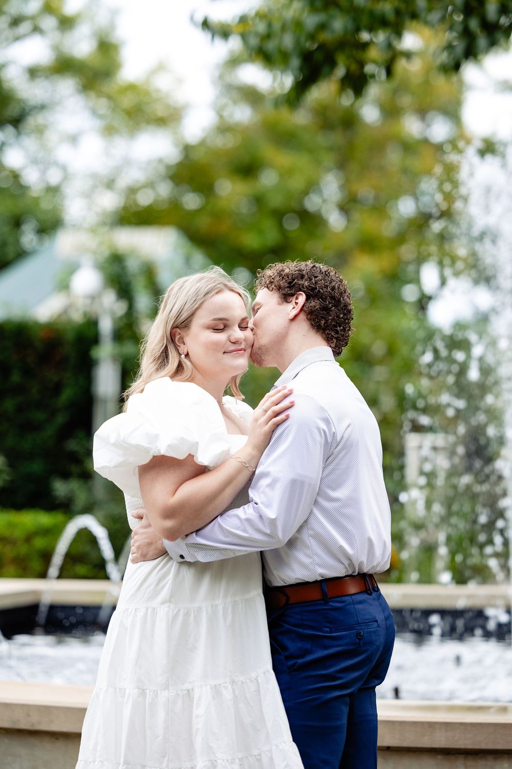 Romantic couple embracing near a garden fountain, man kissing woman in white ruffled dress among green trees.