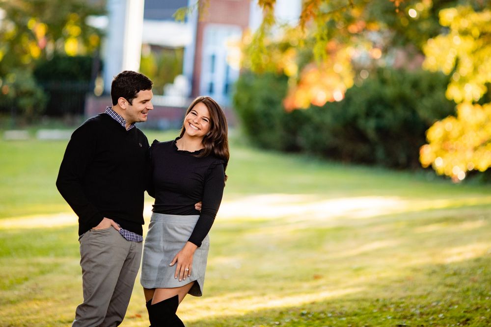 Happy couple walking hand in hand on a sunny lawn surrounded by golden fall trees during engagement photos.