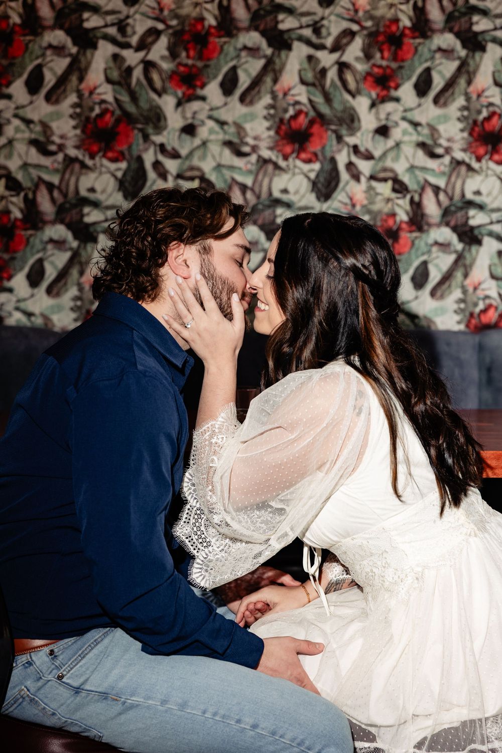 Couple shares an intimate almost-kiss while seated together in front of a floral patterned backdrop.