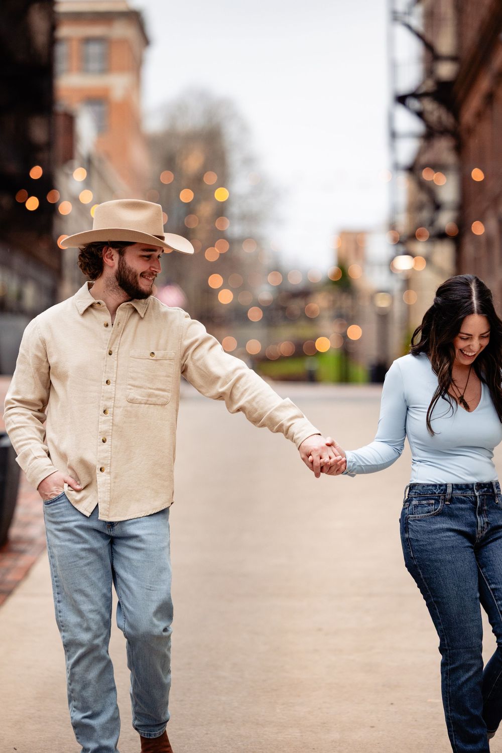 Couple walks hand in hand down a charming city street, man wearing a cowboy hat under string lights.