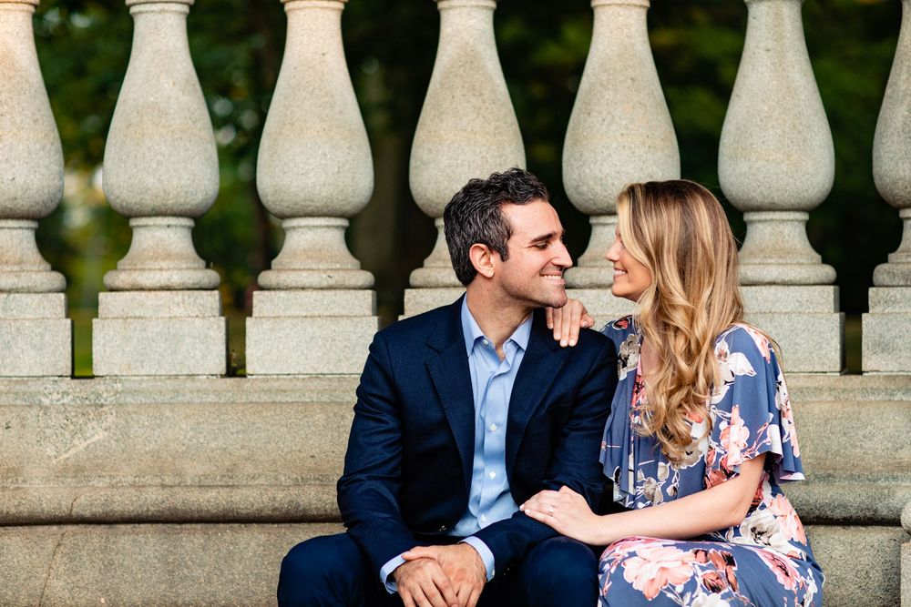 Couple sitting closely together on stone steps near decorative columns, smiling at each other affectionately.