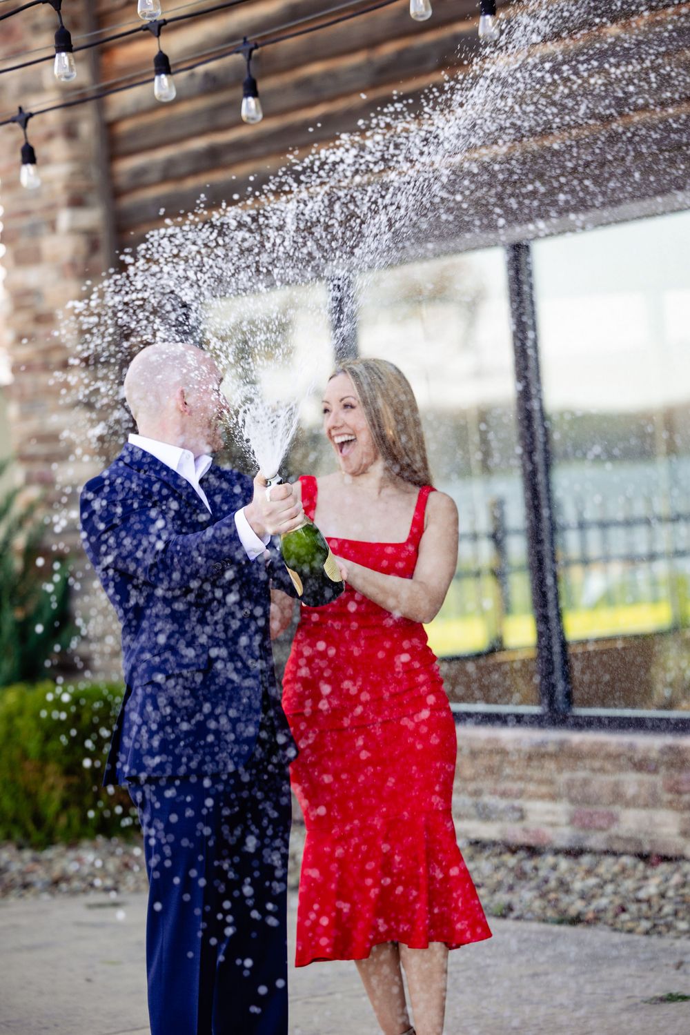 Joyful couple popping champagne bottle outdoors, woman in red dress laughing as sparkling wine sprays everywhere.