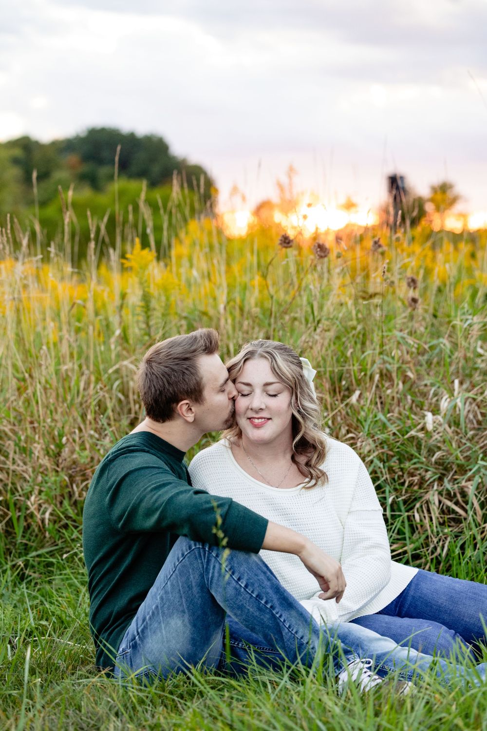 Couple sitting in a meadow at golden hour sunset, man kissing womans cheek as she smiles peacefully.