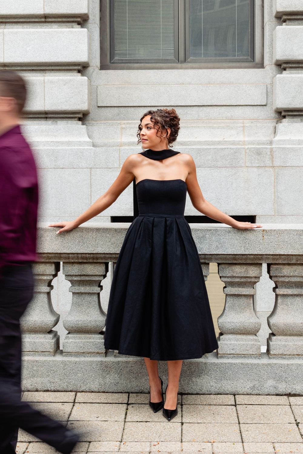 Woman in elegant black strapless midi dress with choker poses gracefully against stone balustrade.