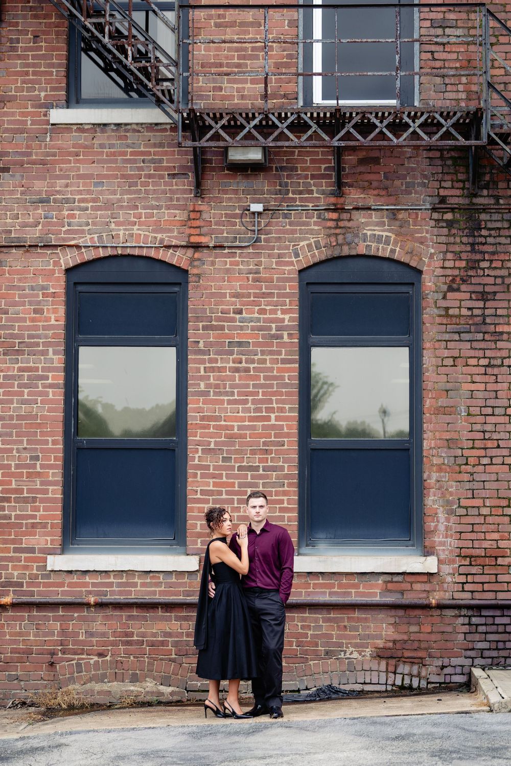Couple posing romantically in front of a rustic brick building with arched windows and a fire escape.