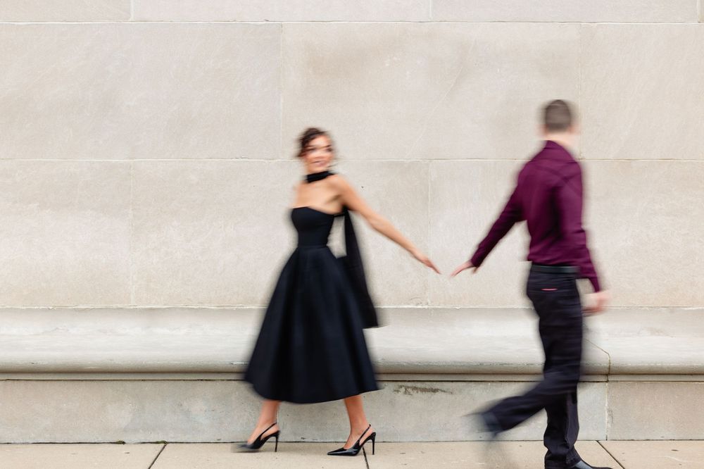 A woman in a black dress and heels holds hands with a man in a burgundy shirt, captured in motion blur.
