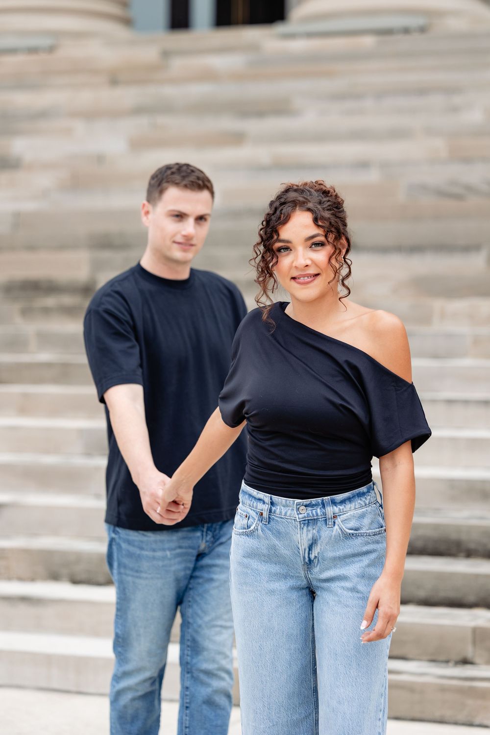A couple in matching black tops and jeans hold hands on stone steps, woman in off-shoulder top smiles at camera.
