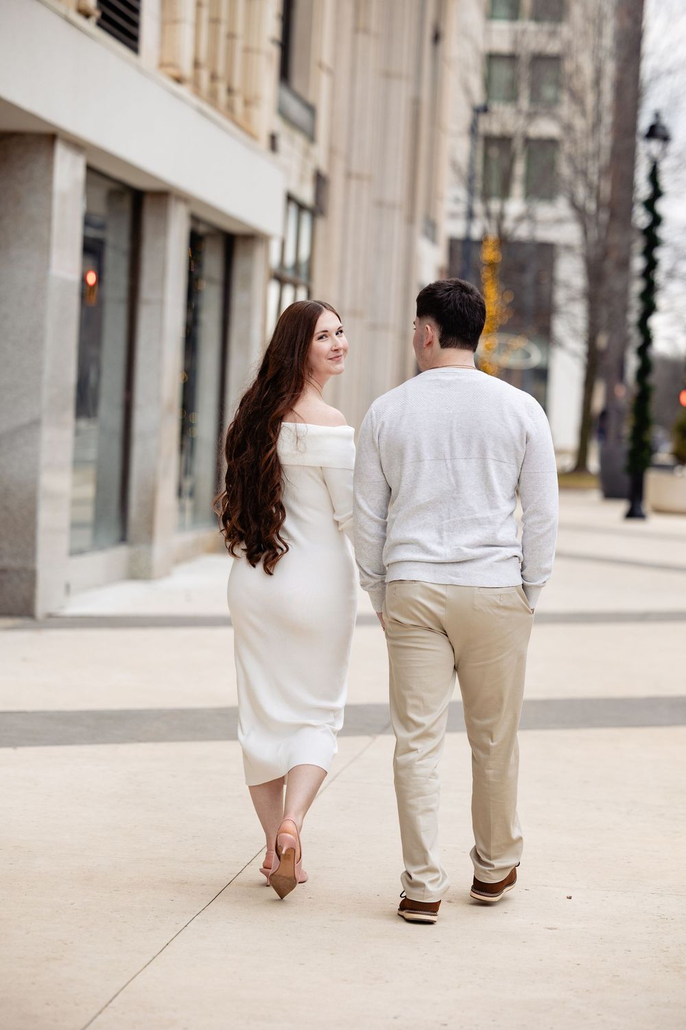 Couple walks away down an urban sidewalk, woman in white dress glancing back over her shoulder.