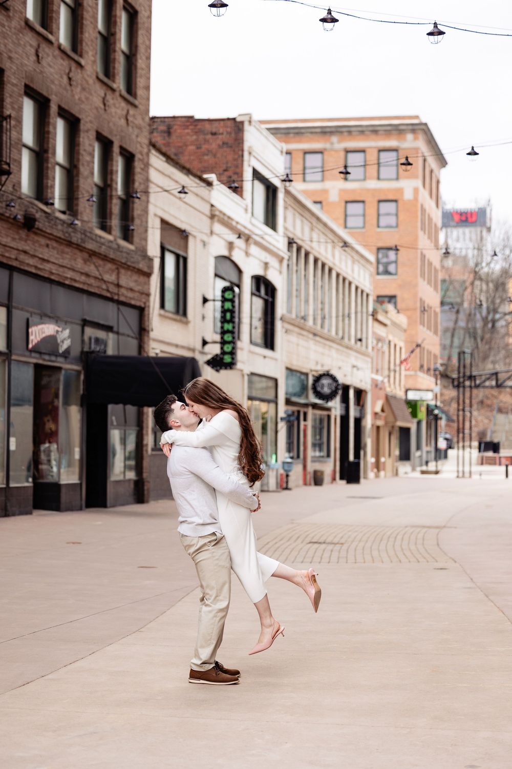 Man lifts and spins his partner joyfully on a quiet downtown city street during engagement photos.