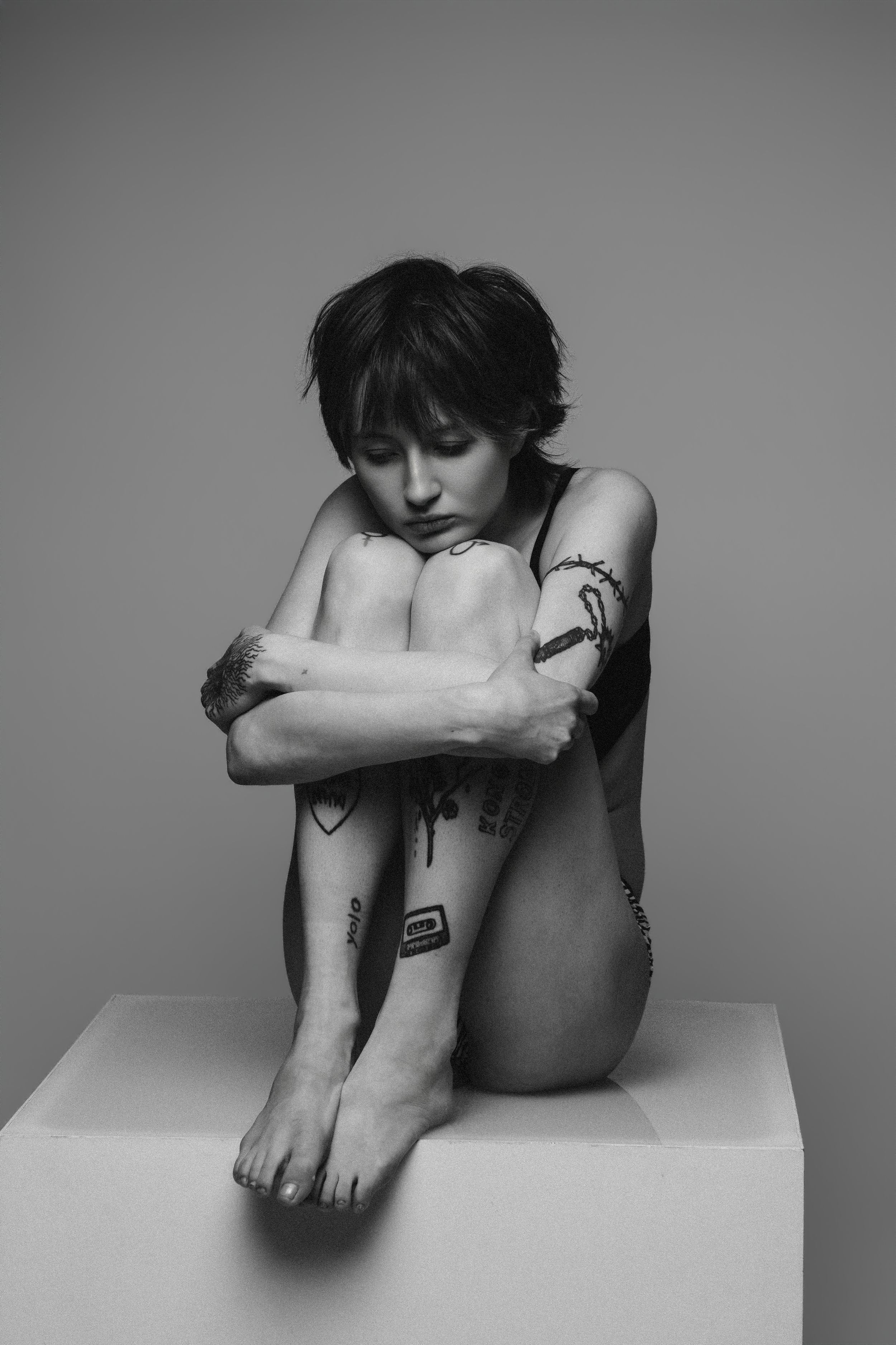 Black and white artistic nude photograph showing a person in a curled pose with tattoos sitting on a white surface.