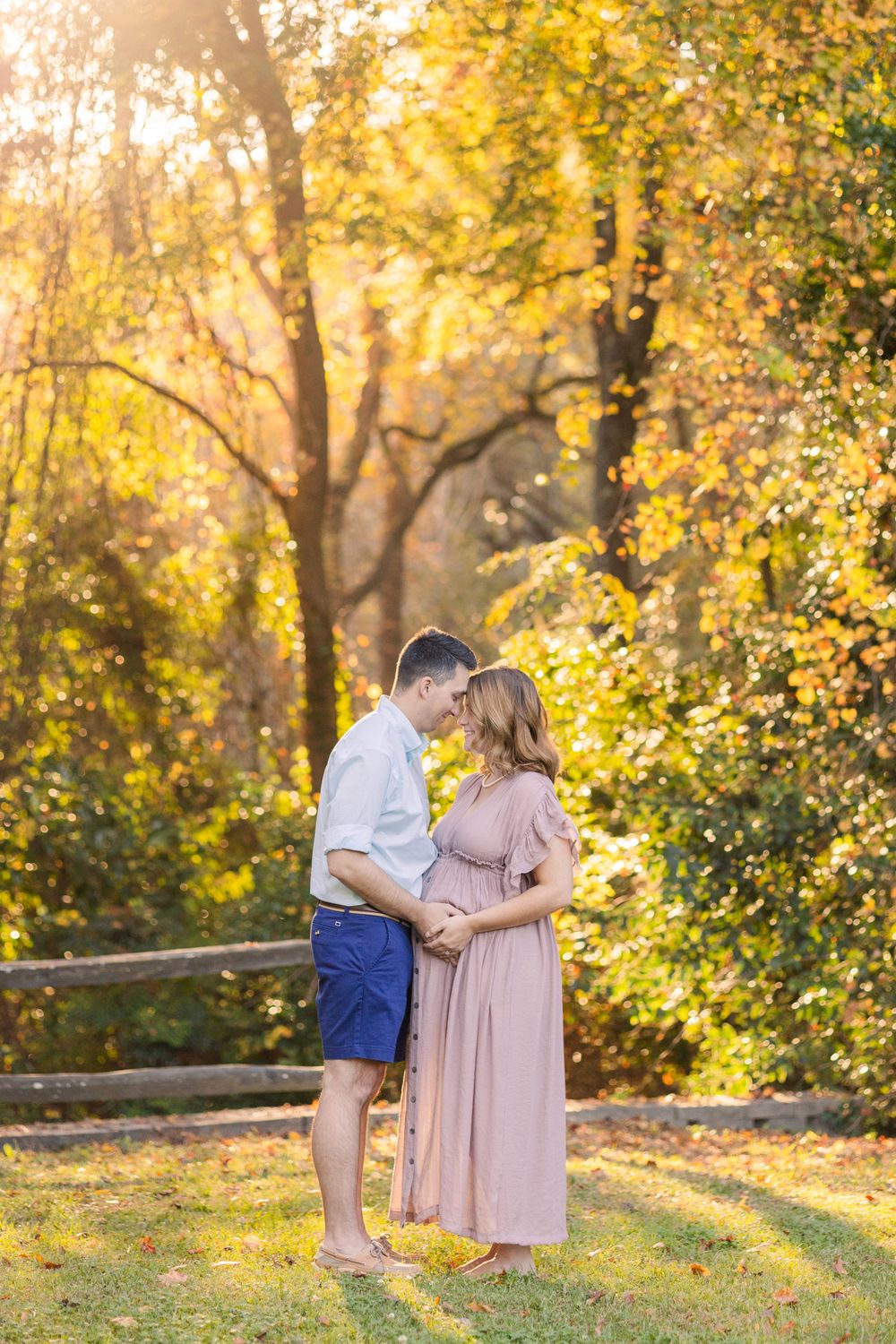 A romantic sunset engagement photo featuring a couple in pastel outfits standing together among golden autumn trees.
