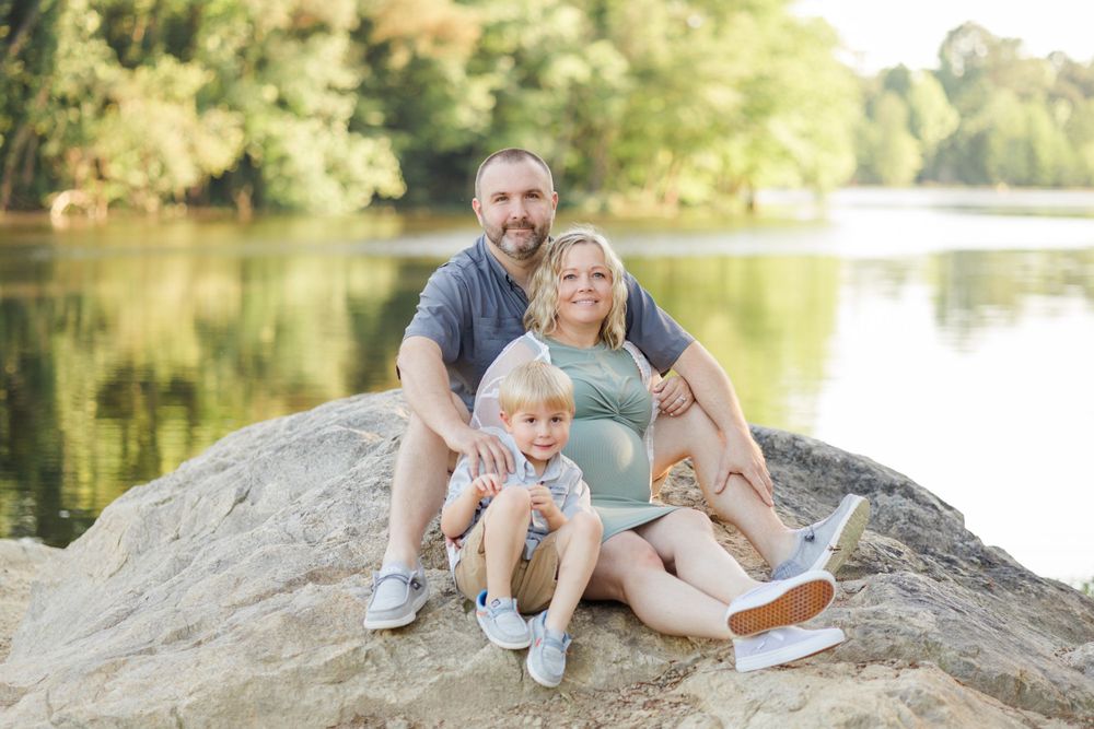 Family sitting together on large rock by peaceful lake during golden hour.