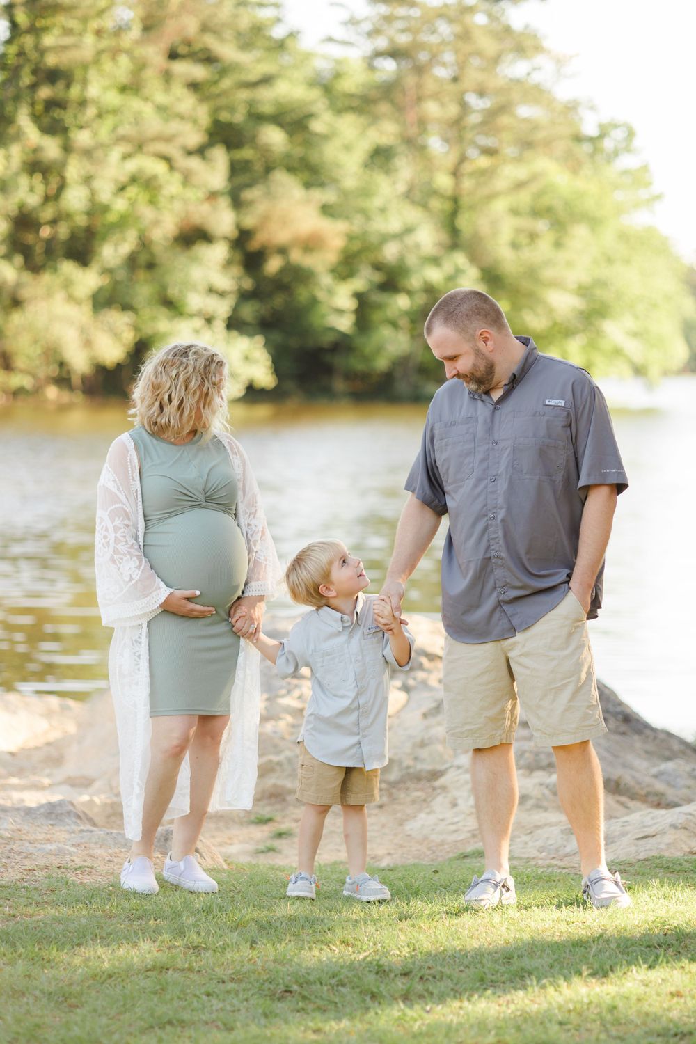 Family of three posing together by a scenic lake during sunset.
