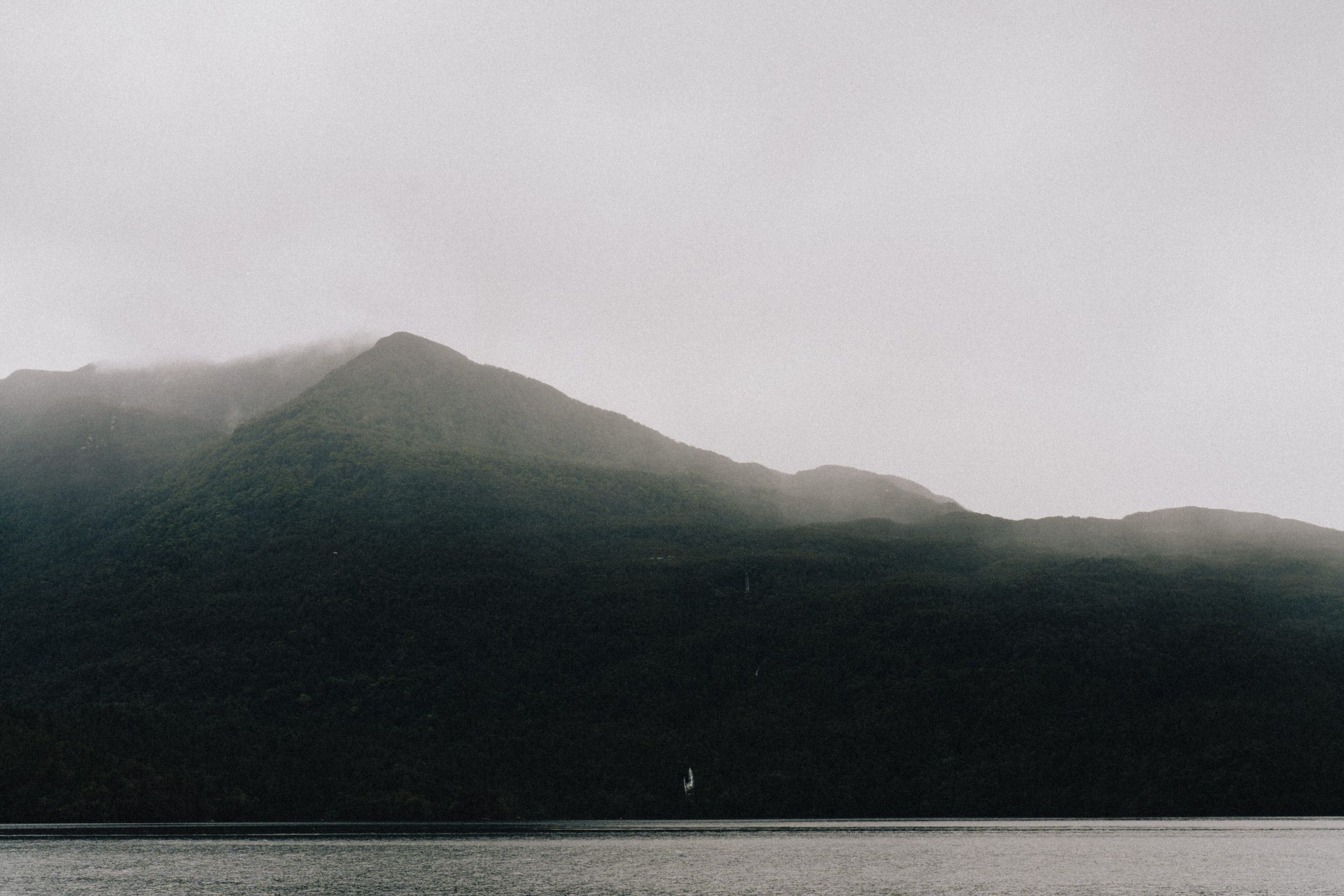 Misty mountain silhouette rises above dark waters under overcast sky.