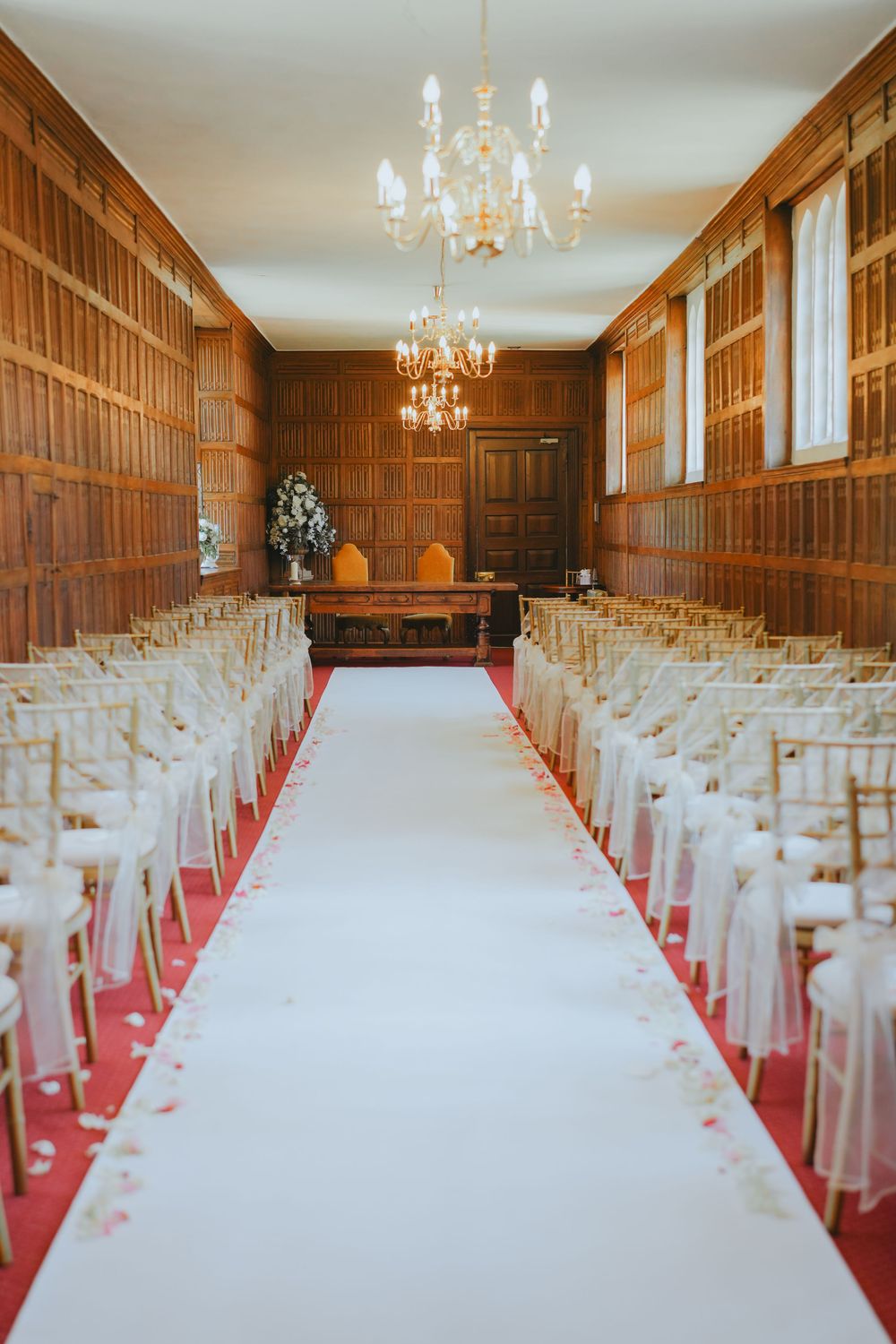 Elegant wood-paneled wedding ceremony room with white aisle runner, covered chairs and crystal chandeliers.