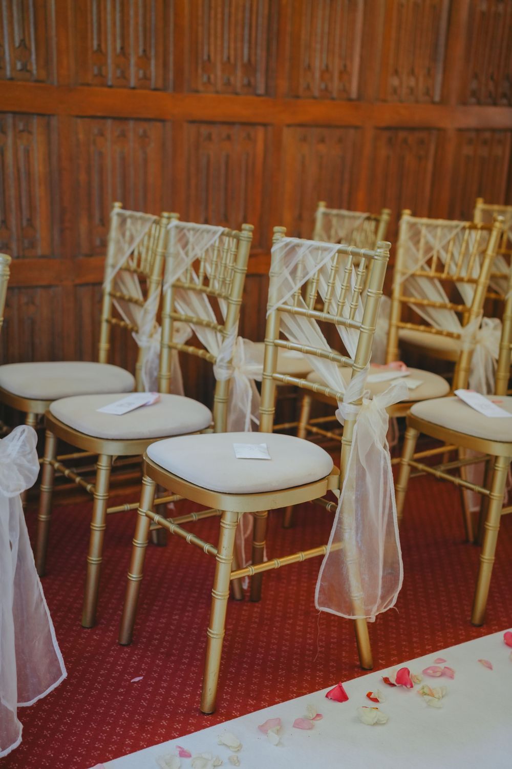 Elegant gold chiavari chairs adorned with white organza sashes stand in rows on a red carpet at a wedding ceremony setup.