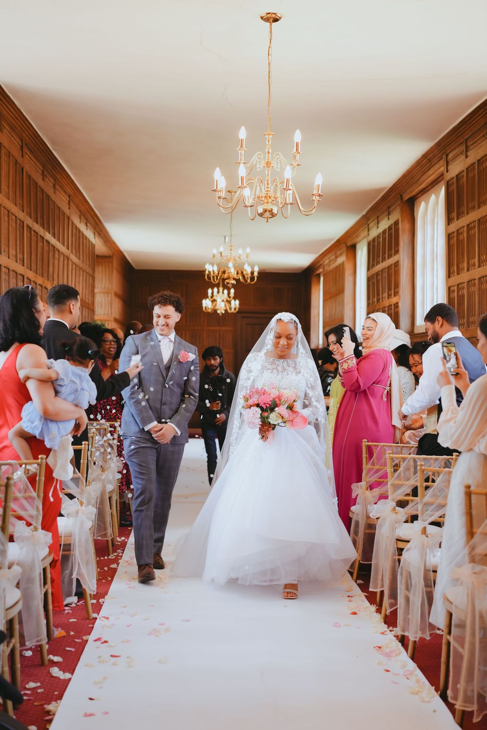 Elegant indoor wedding ceremony with guests lining a red-carpeted aisle under crystal chandeliers.
