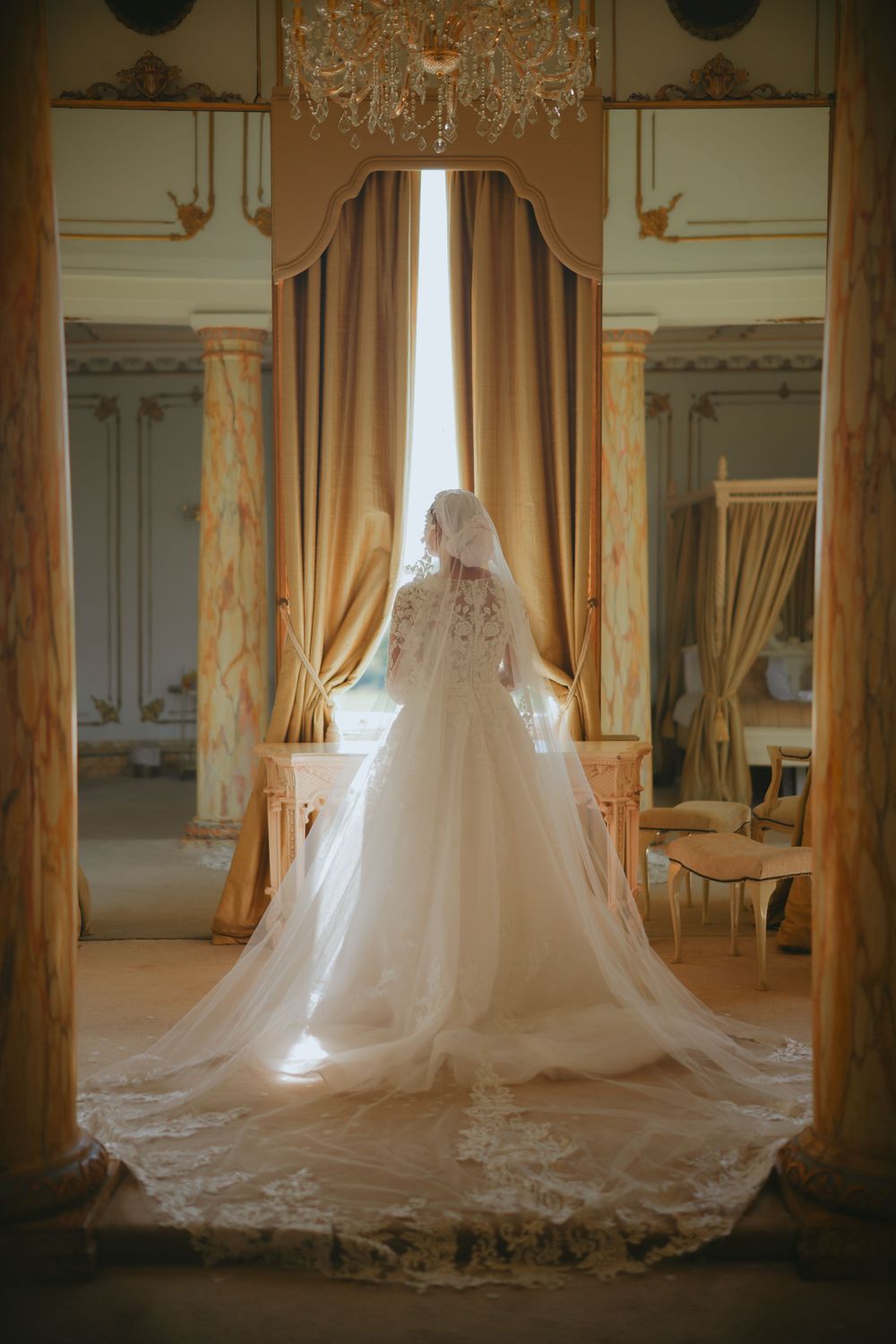 A stunning white lace wedding gown with a long flowing train stands elegantly in an ornate room with golden drapes.