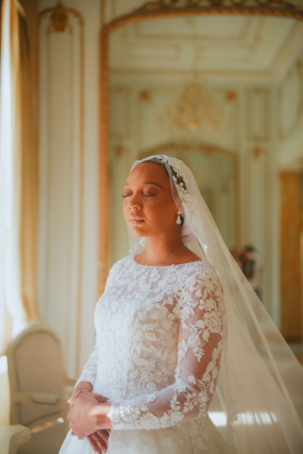 Elegant bride in white lace long sleeve wedding dress and veil stands in ornate room with natural sunlight.