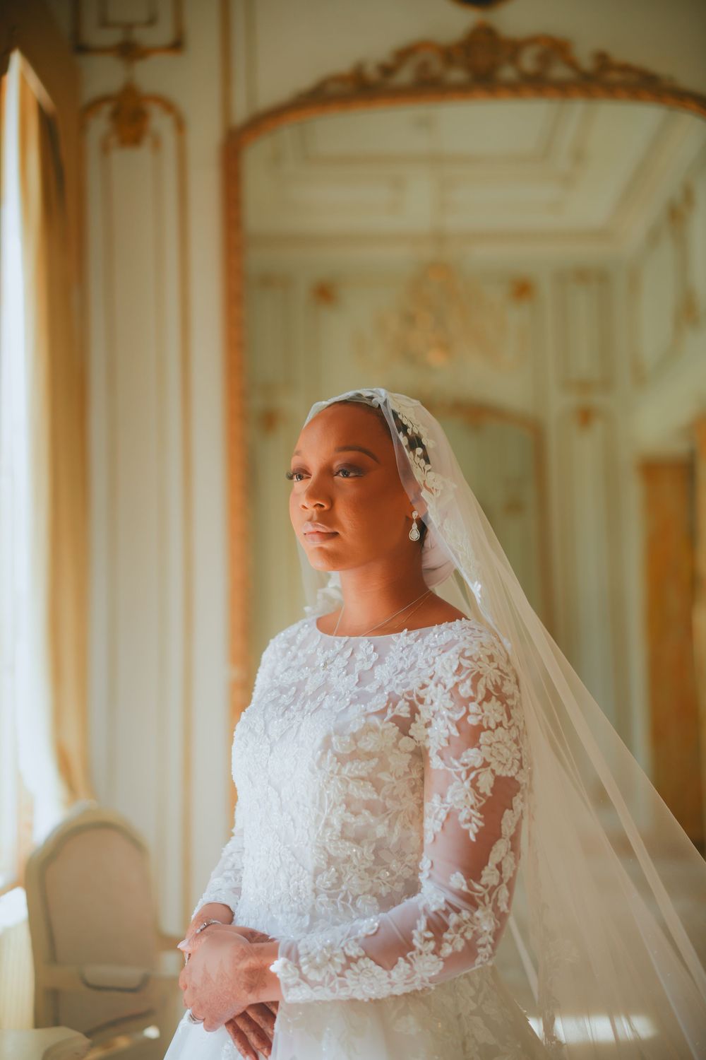 Bride in elegant white lace wedding dress with long sleeves and flowing veil poses in ornate room with golden decor.