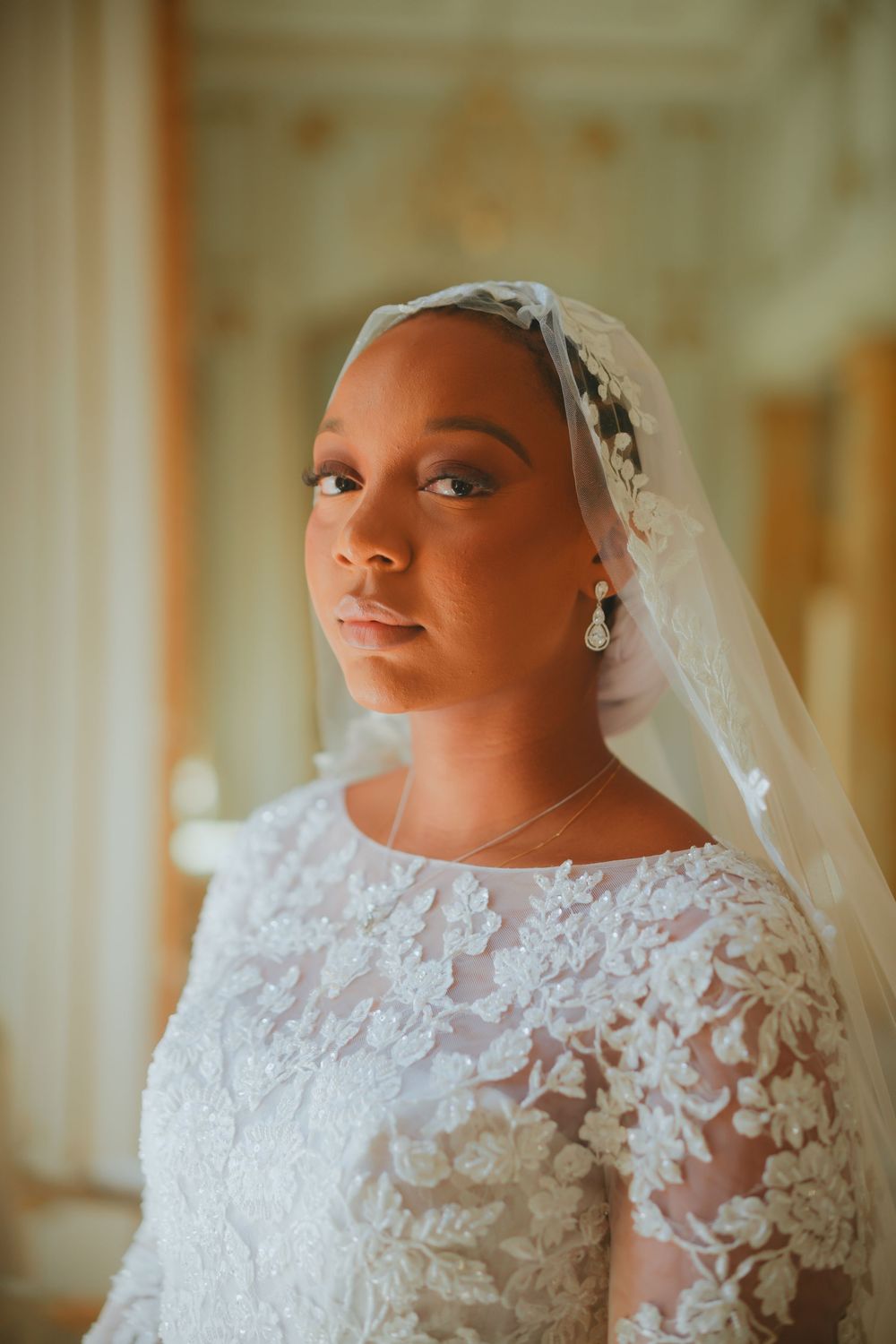 A bride wearing an elegant white lace long-sleeve wedding gown and delicate veil poses in a warmly lit room.