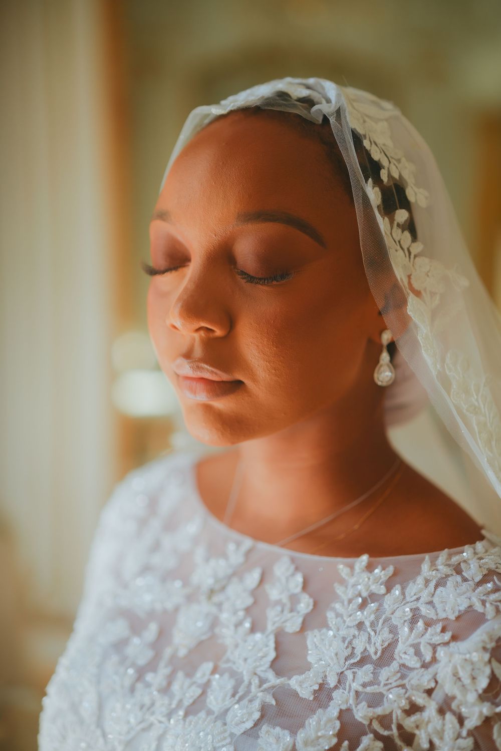 A bride wearing a delicate lace wedding dress and floral headpiece with veil poses with eyes closed in soft lighting.