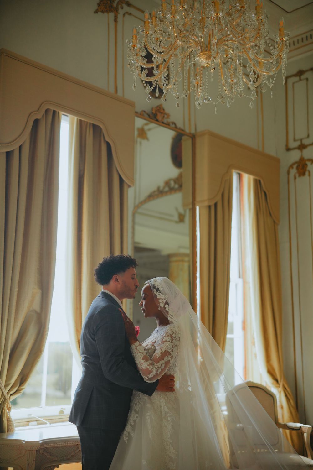 A wedding couple dances together in an ornate venue with vintage drapes and crystal lighting.