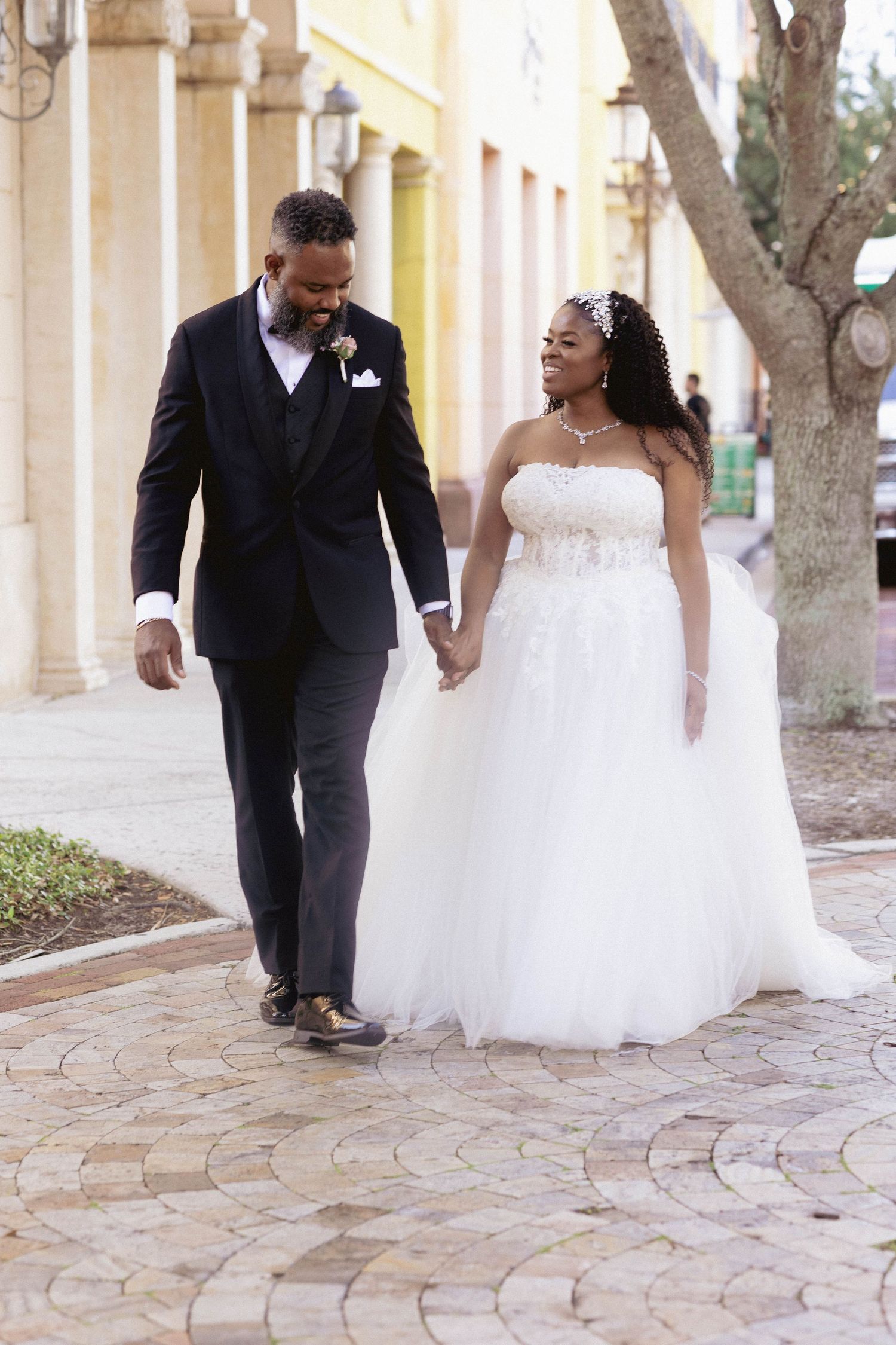 Newlywed couple walks hand in hand in wedding attire along colorful outdoor courtyard.