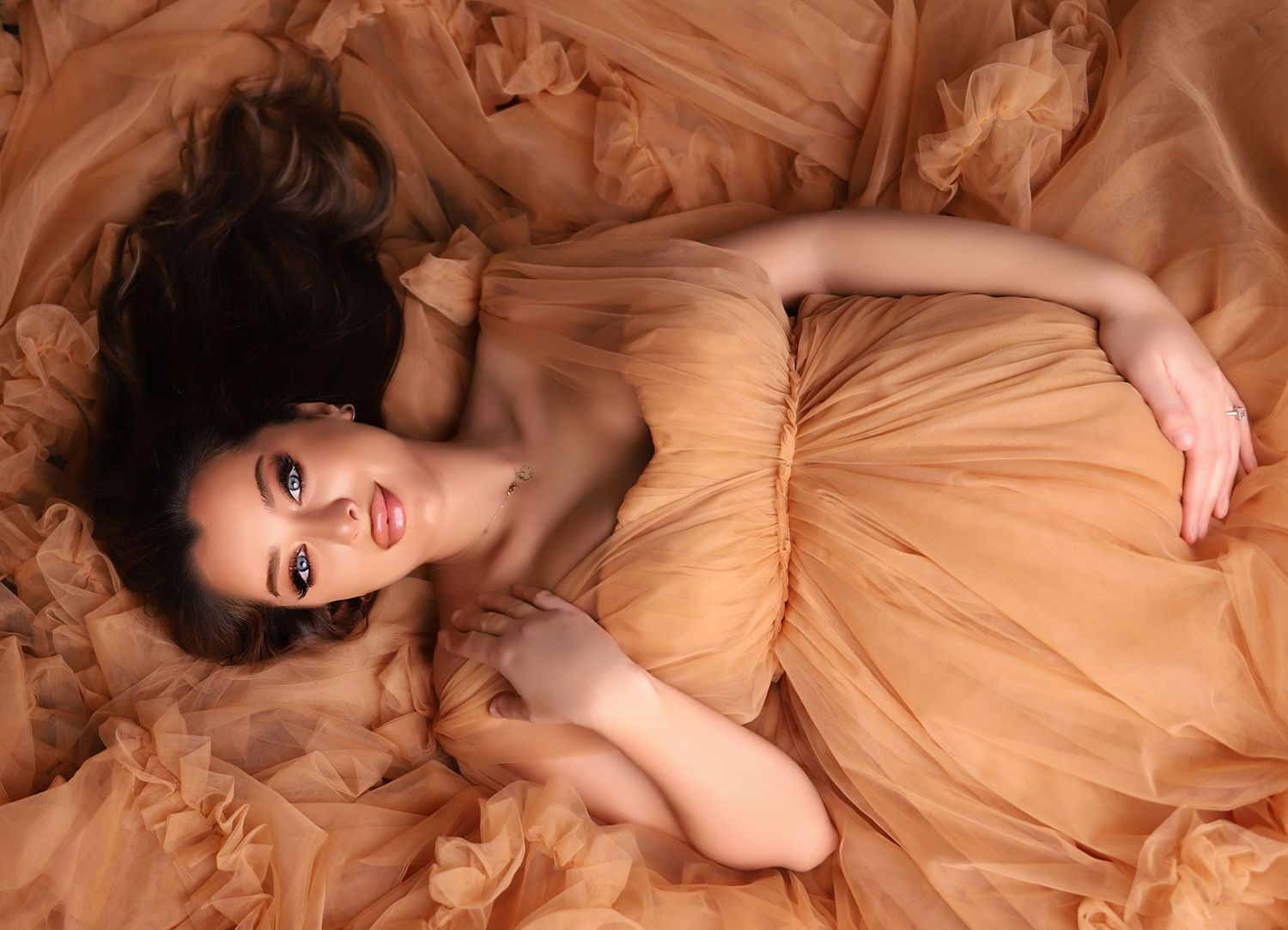 Woman in flowing peach gown poses elegantly on floor in artistic maternity photos.
