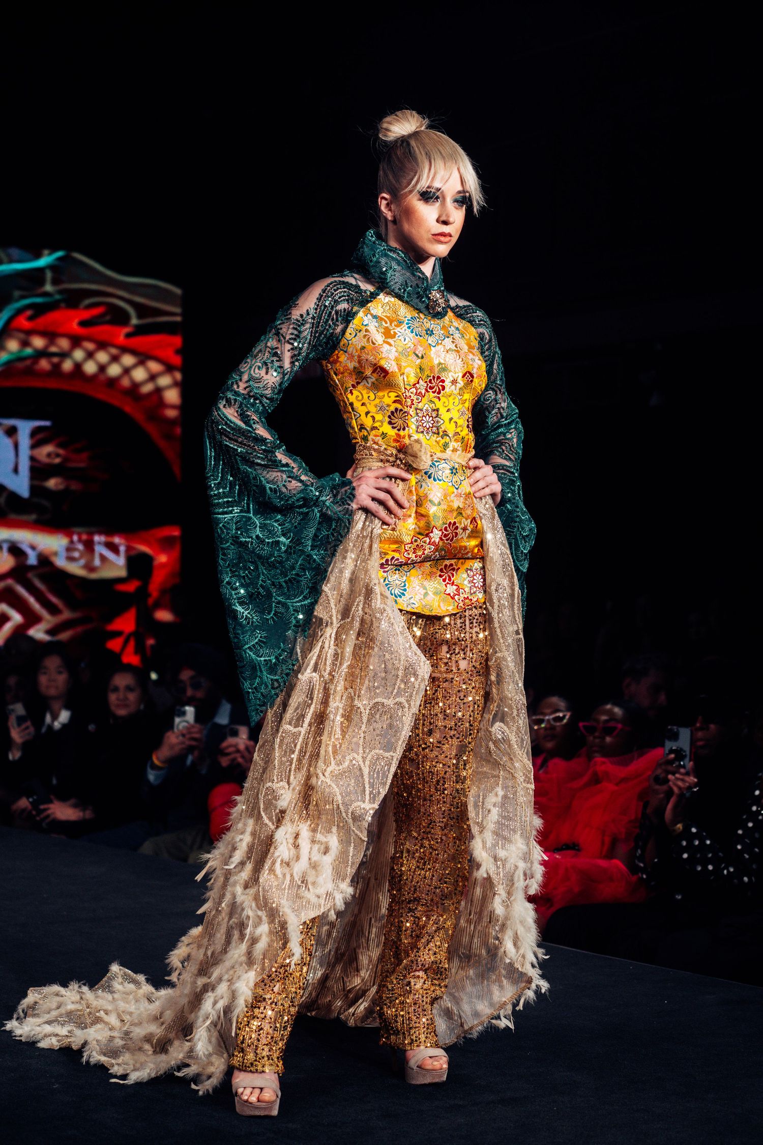 A fashion model walks the runway wearing a shimmering gold and turquoise dress with flowing transparent cape details.