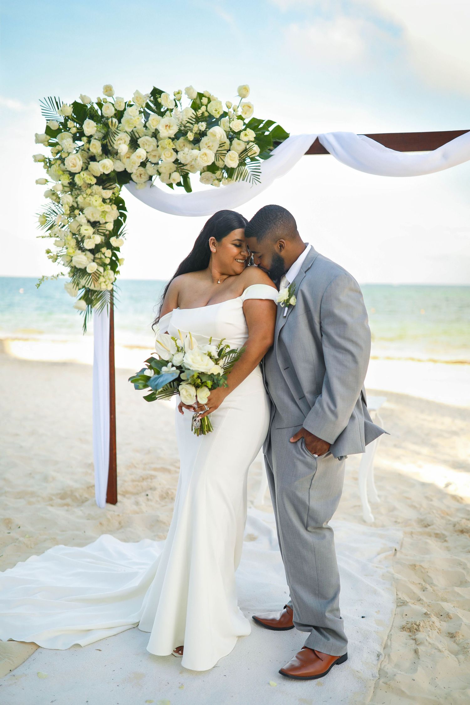 Beach wedding ceremony setup with white floral arch and couple sharing intimate moment on sandy shore.