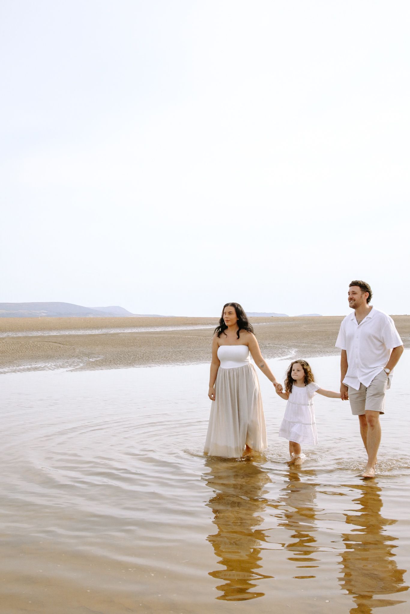 Family in white outfits holding hands while walking together through shallow ocean waters at sunset.