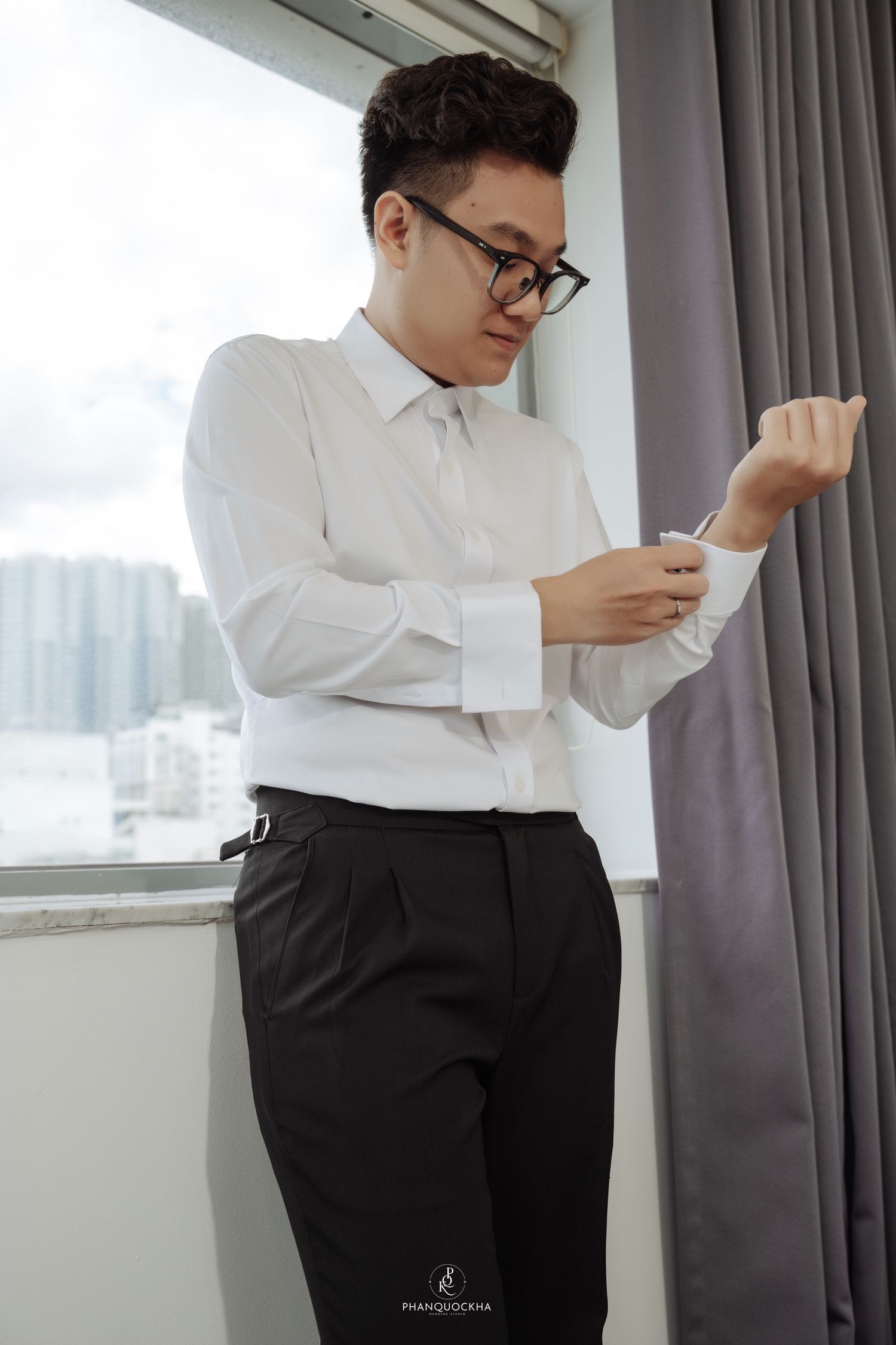 Asian groom in white shirt and black trousers buttoning his cuff near a bright window.
