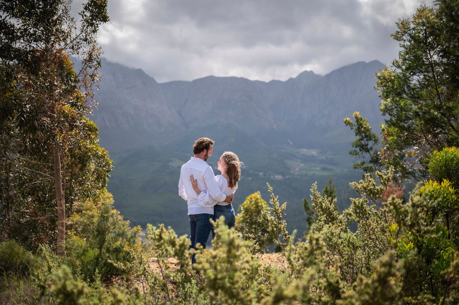A couple enjoys a scenic mountain view while standing among lush greenery on a cloudy day.