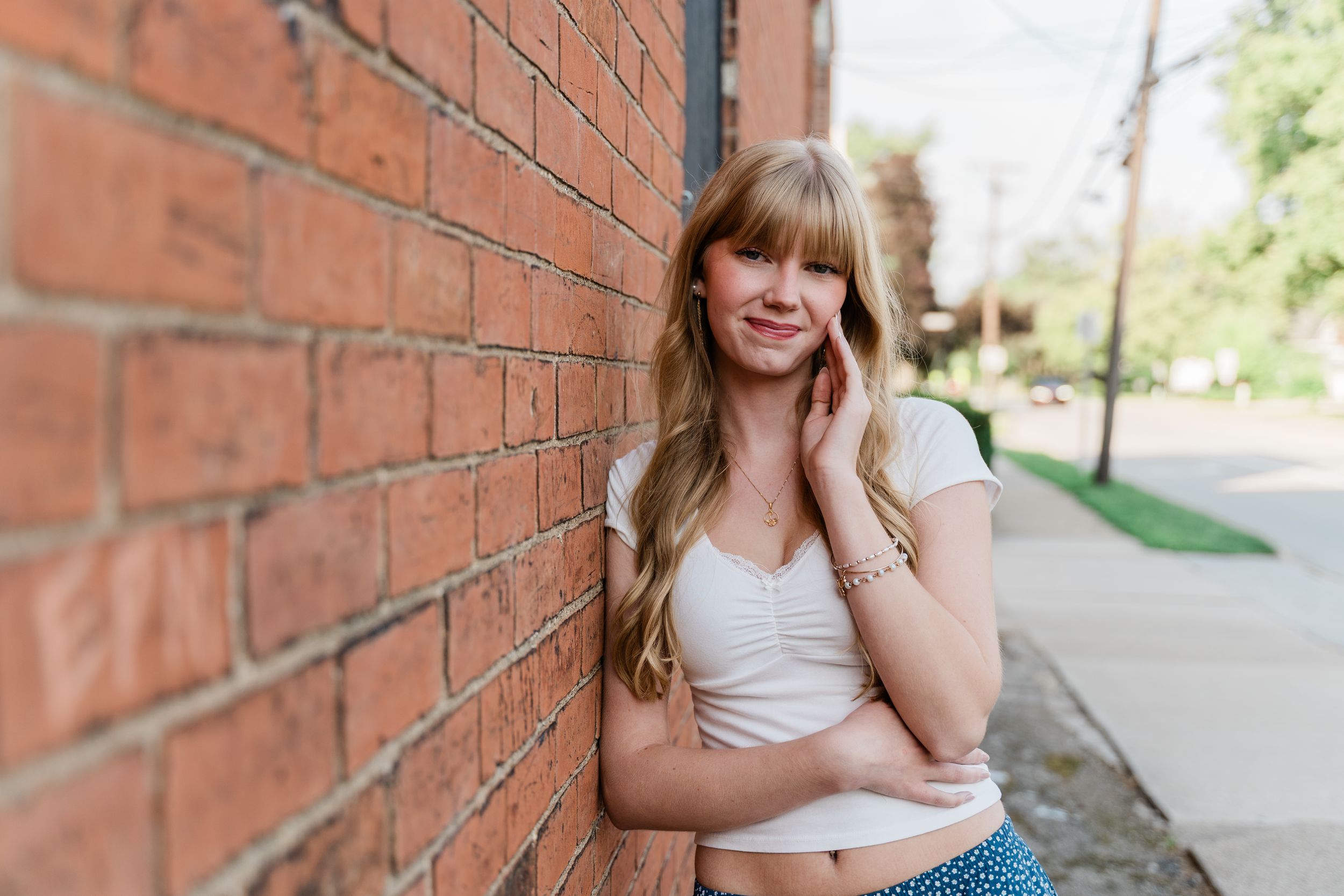 A person in a white top and patterned skirt leans casually against a red brick wall outdoors on a sunny day.