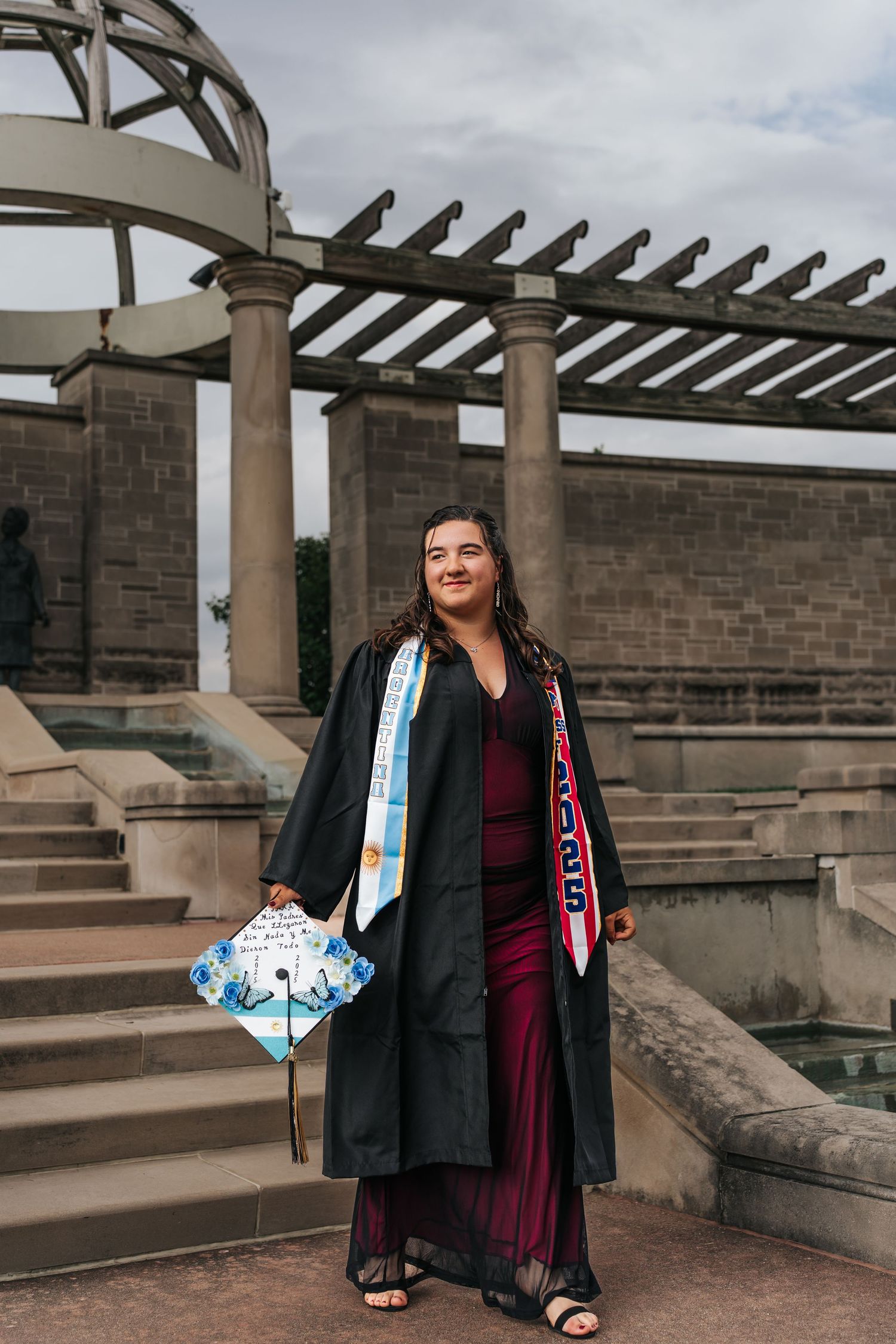 Graduate in burgundy dress and academic regalia stands proudly on stone steps beneath pergola.