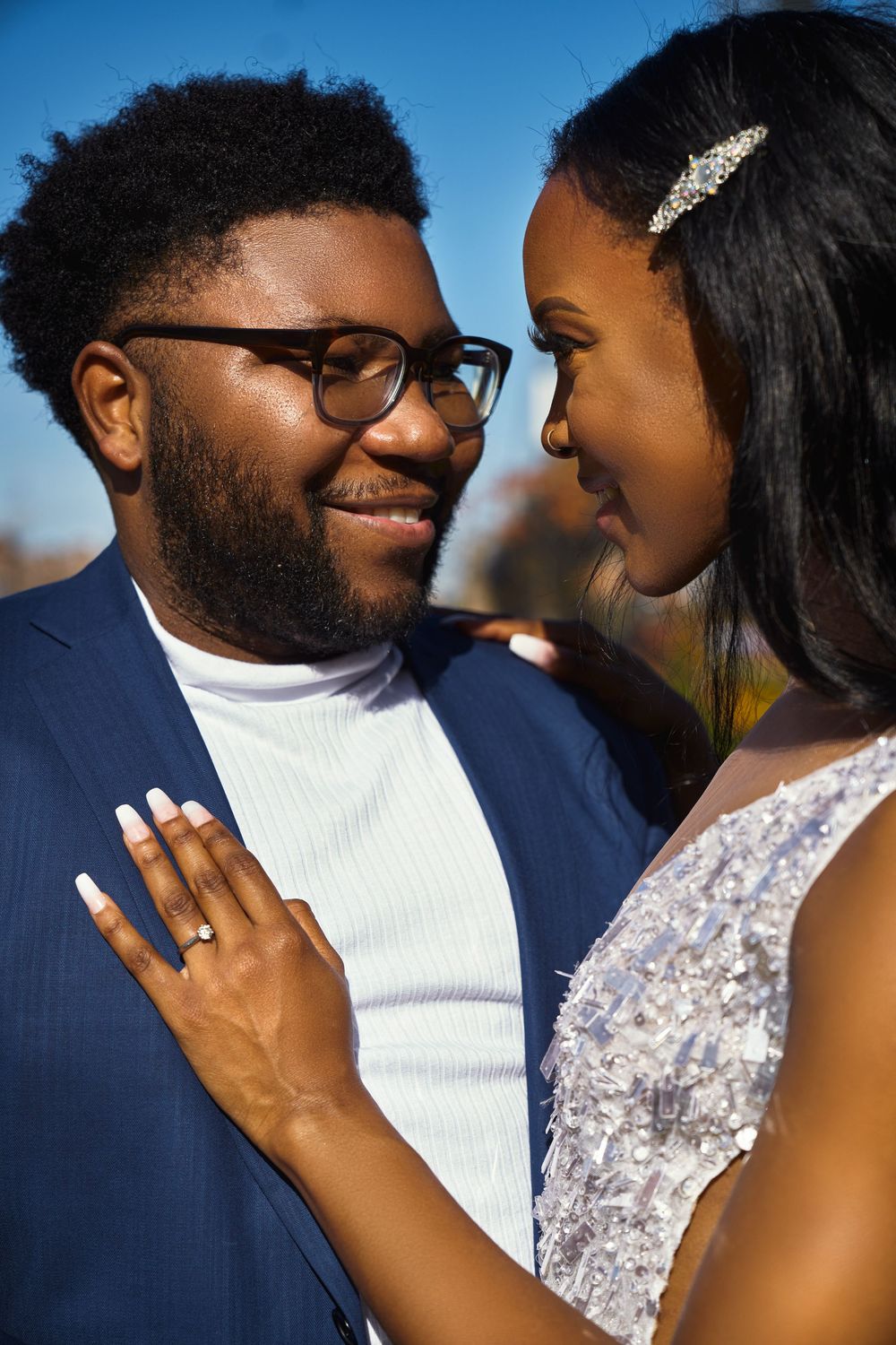 Romantic couple in wedding attire share an intimate moment against blue sky as bride displays engagement ring at Brooklyn Bridge