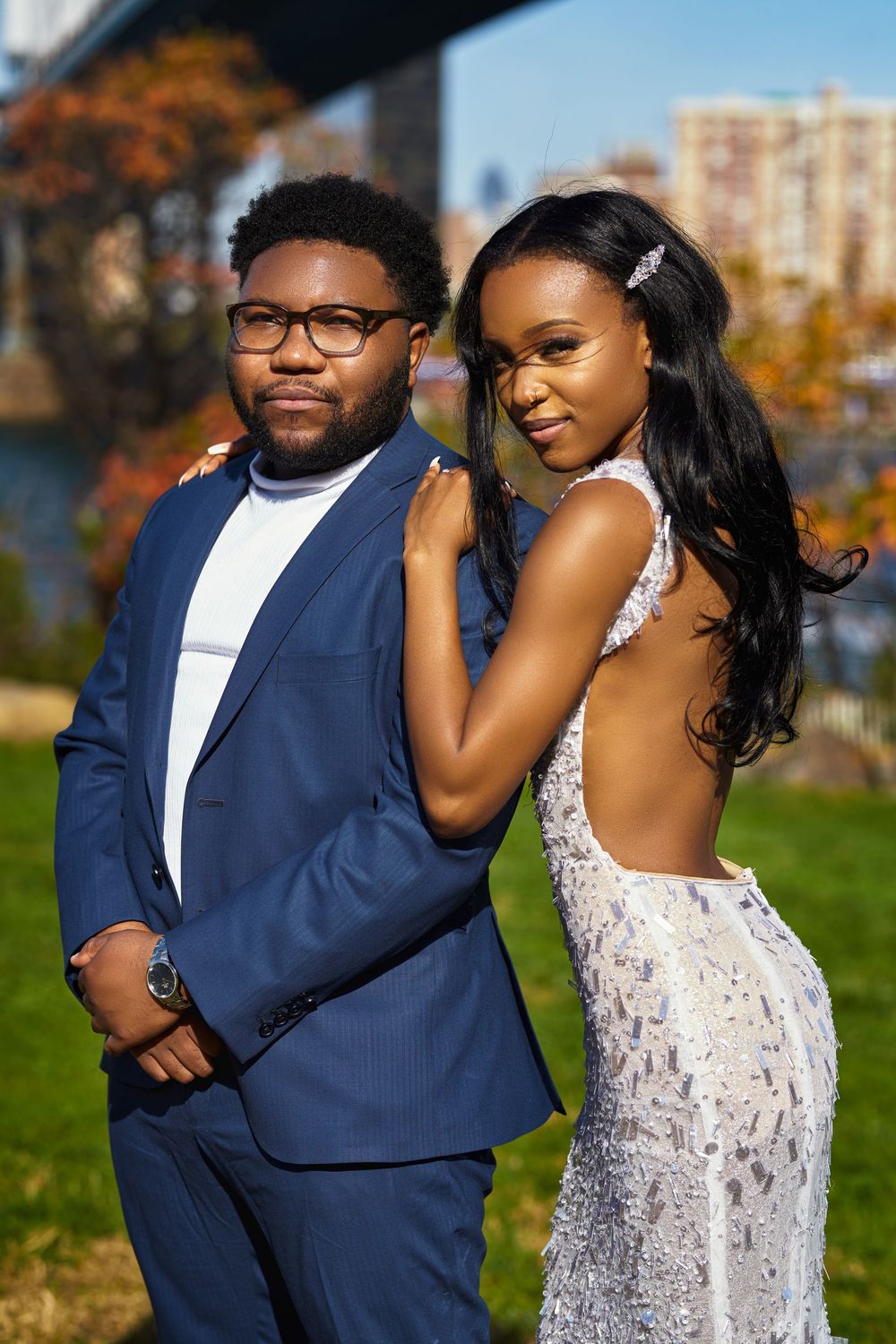 A well-dressed couple poses together outdoors, with one wearing a blue suit and the other in an elegant white lace gown at Brooklyn Bridge
