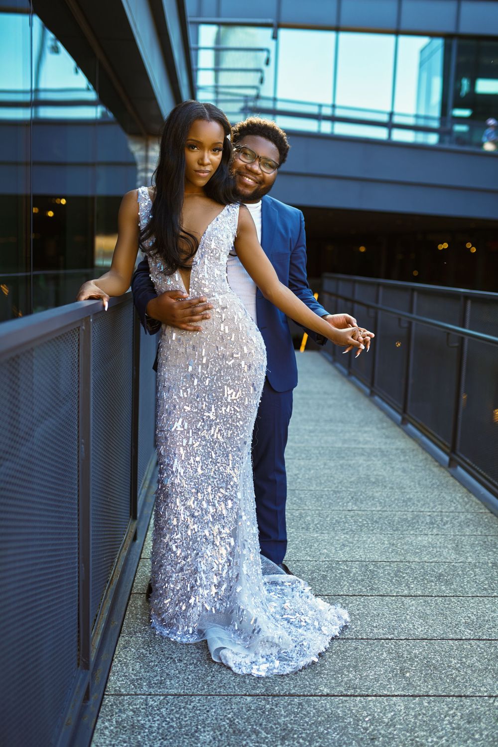 A couple in elegant attire pose together on a modern walkway, with her in a sequined white gown and him in a navy suit at Brooklyn Bridge