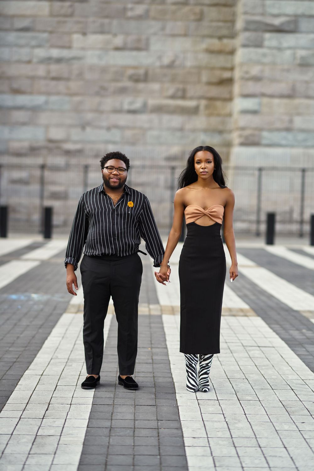Stylishly dressed couple holding hands while standing on patterned pavement near stone wall at Brooklyn Bridge