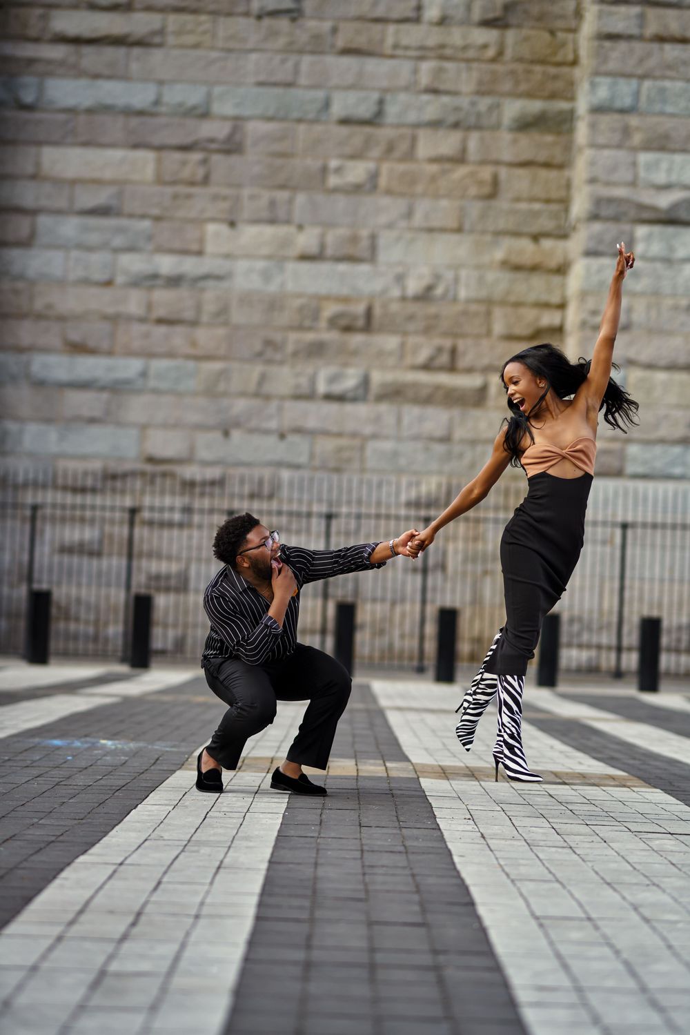 Two dancers perform a dynamic urban dance move together on a paved plaza with stone walls in the background at Brooklyn Bridge